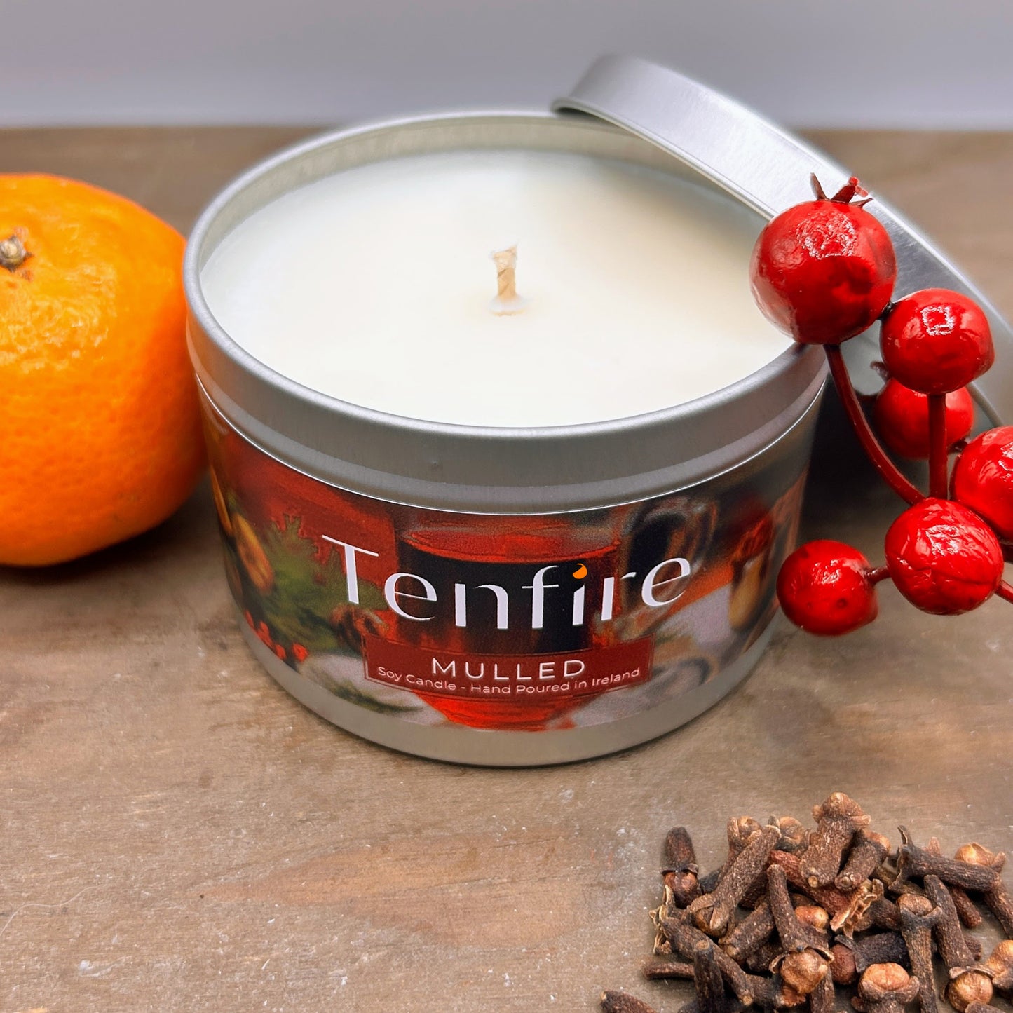Tenfire mulled candle in a tin with an orange, red berries, and cloves on a wooden surface.