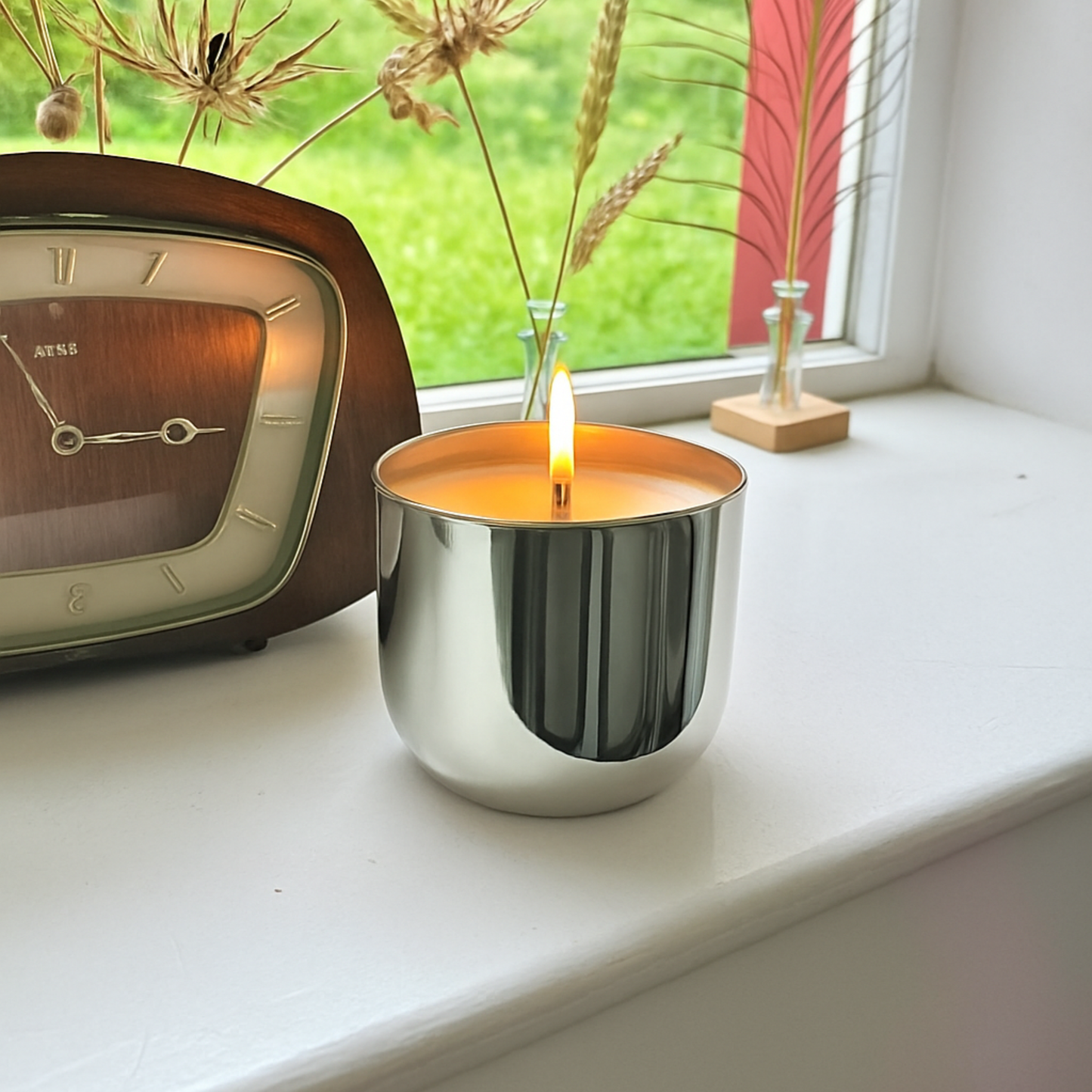 Smooth silver candle vessel on a windowsill in front of a clock and window
