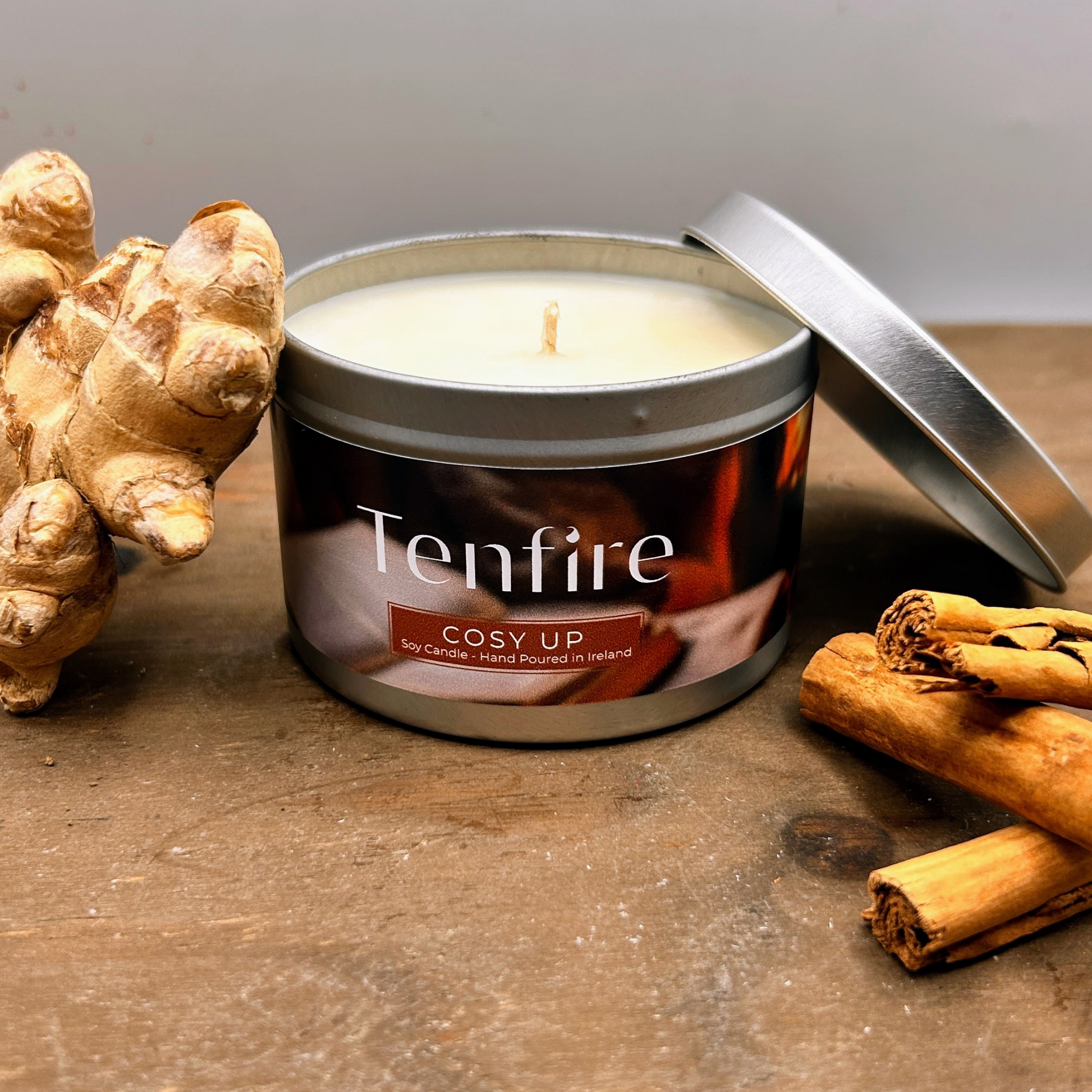 open candle tin called cosy up sitting on a wood board with ginger and cinnamon by the side