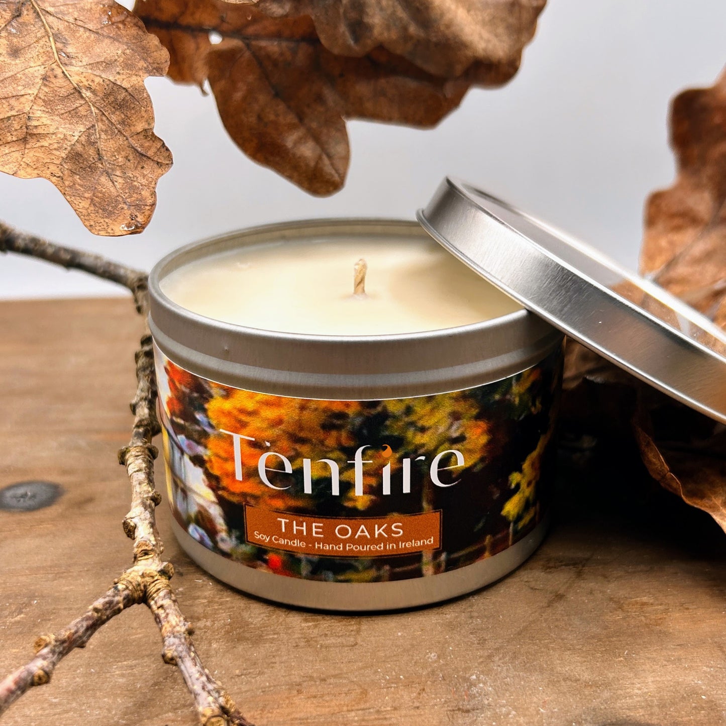 Candle in a metal tin labeled 'Tenfire The Oaks' surrounded by autumn leaves and branches.