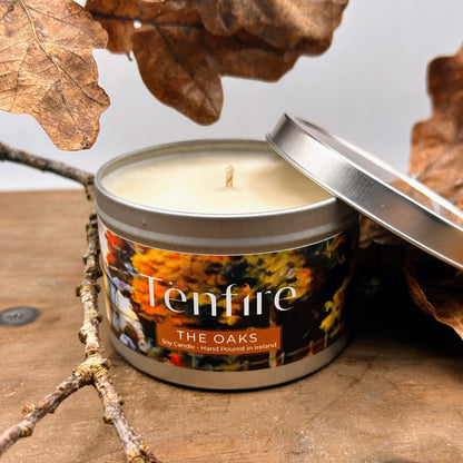 Candle in a metal tin labeled 'Tenfire The Oaks' surrounded by autumn leaves and branches.